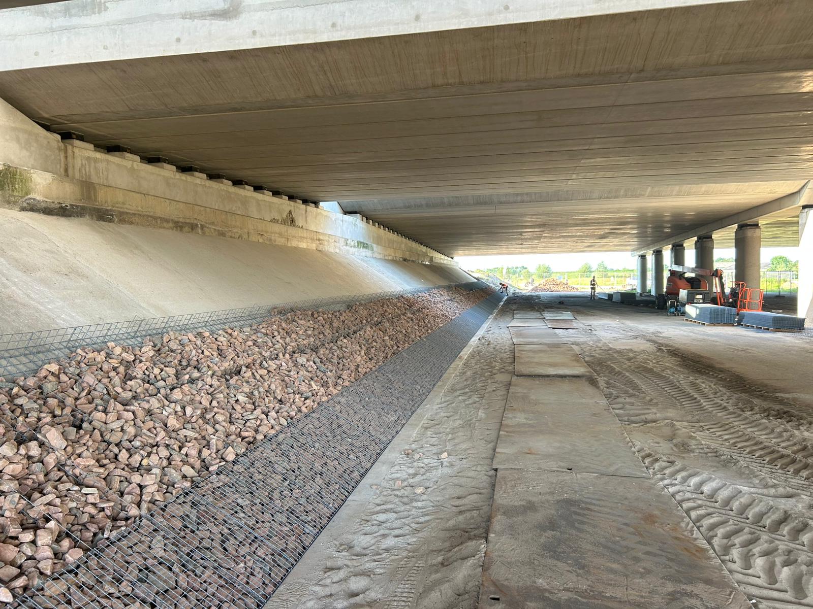 Grondkering met steenkorven onder een snelwegviaduct, gevuld met rode breuksteen